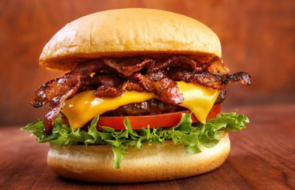 Juicy sandwich with bacon, beef patty, lettuce, tomato, and cheese between two buns on a wooden Table. Keep reading for the full guide to the best burger spots in Gainesville.  