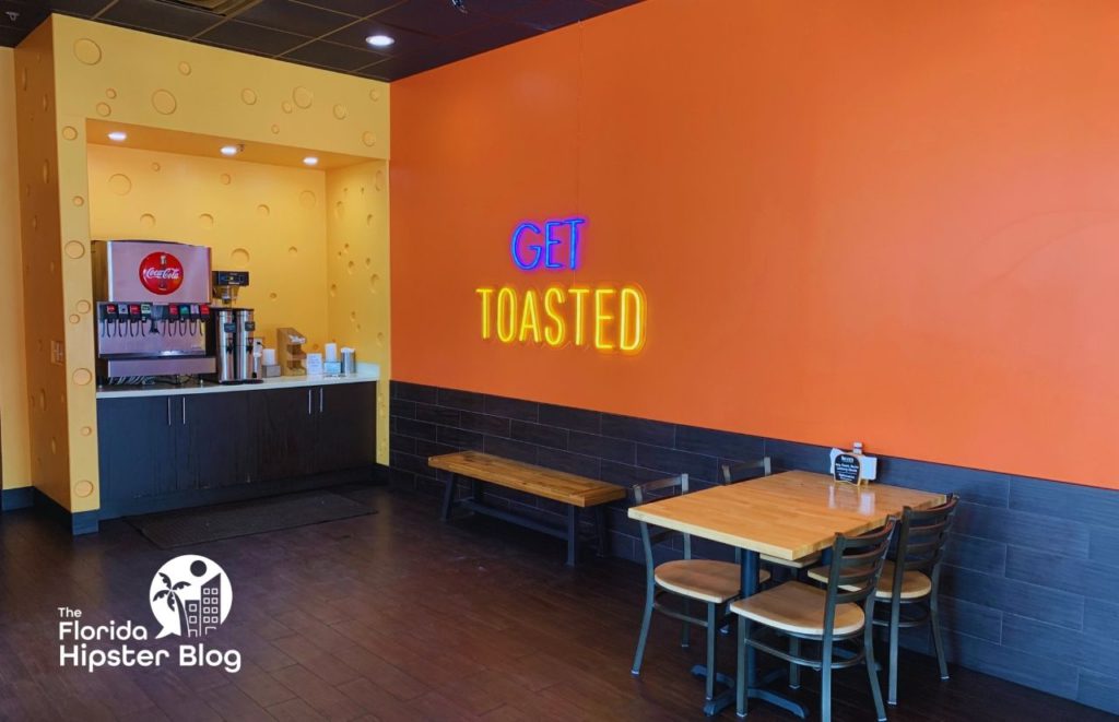 Toasted Interior in Orlando Florida. Get Toasted light up sign sits on orange wall near drink machine. Drink machine wall looks like cheese. Keep reading to see what are the best places to get lunch in Orlando.