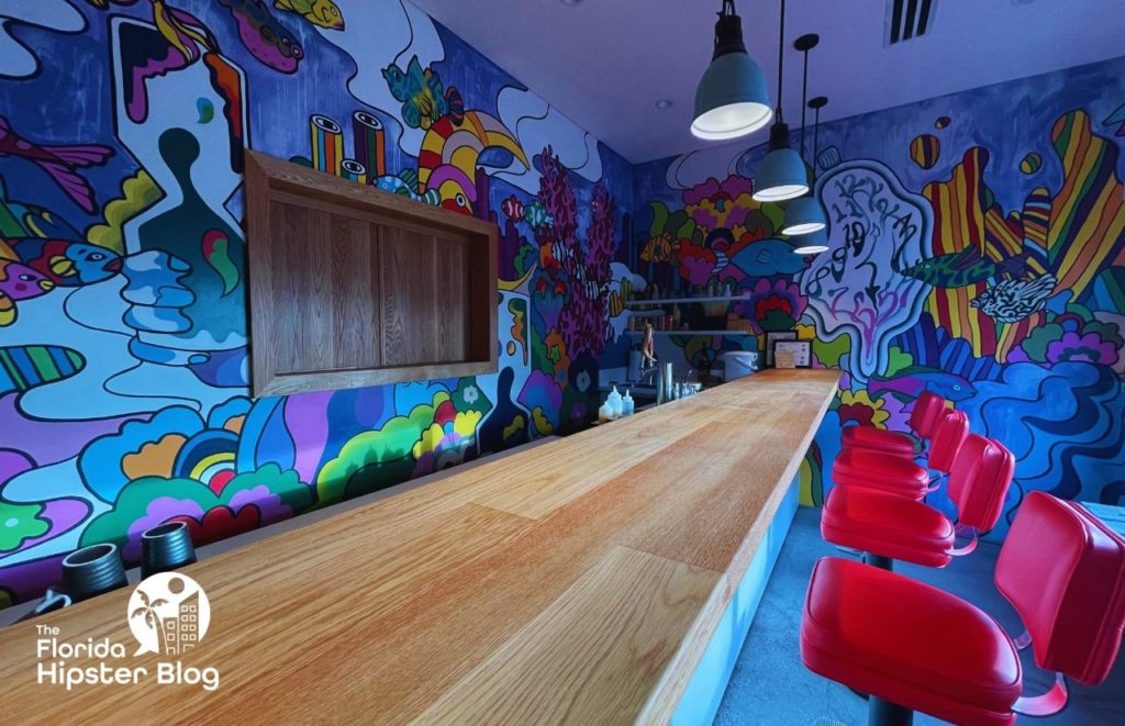 The waiting room at Kadence Sushi in Orlando , Florida features an elaborate mural on the wall, a wood bar, and red bar stools. Keep reading for more on the best restaurants in Orlando, Florida. 