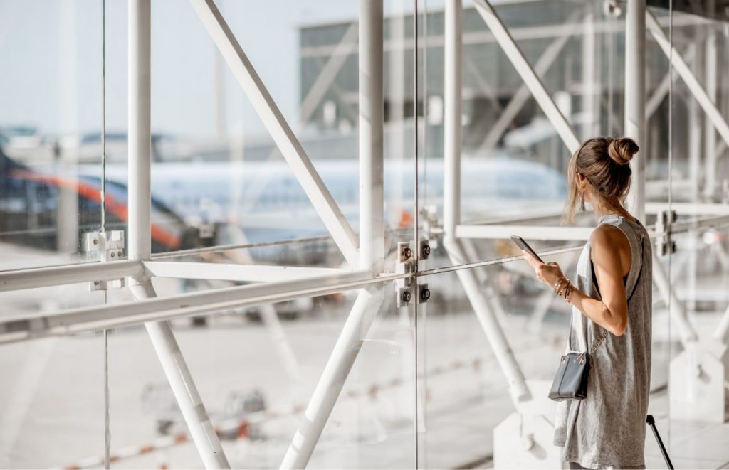 Lady looking out the windows with her personal item and luggage in airport. Keep reading for the full guide to whether or not you can take both a backpack and a carry-on on the plane.  