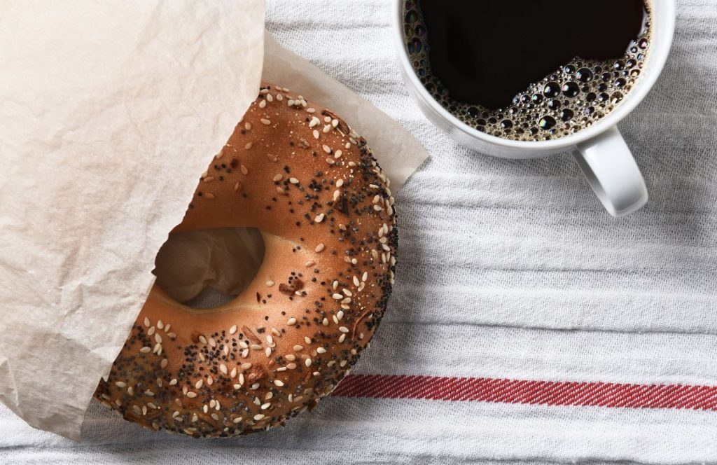 An everything bagel sides out of a paper wrapper onto a table near a cup of coffee. Keep reading to see what are the best places to get breakfast on International Drive in Orlando Florida.