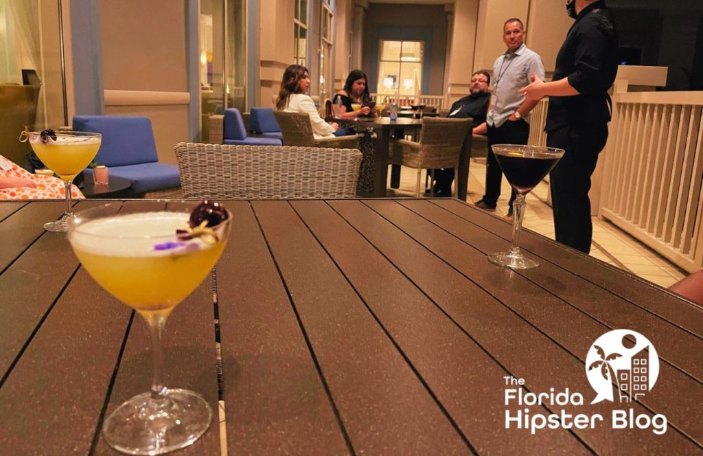 Three martinis sit on a wood table in the Waldorf Astoria in Orlando Florida. People sit in the background talking. Keep reading for more romantic getaways in Orlando.