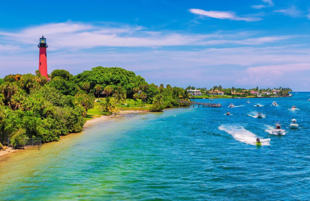 Boats speed along the coast of Jupiter Beach in Florida. The water sits against green trees and a lighthouse. Keep reading for more places to take a perfect day trip from Orlando, Florida. 