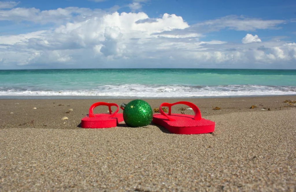 The 12 Days of Christmas on Clearwater Beach with red flip flops next to green ornament on sandy beach. One of the best things to do for Christmas in Clearwater Beach, Florida