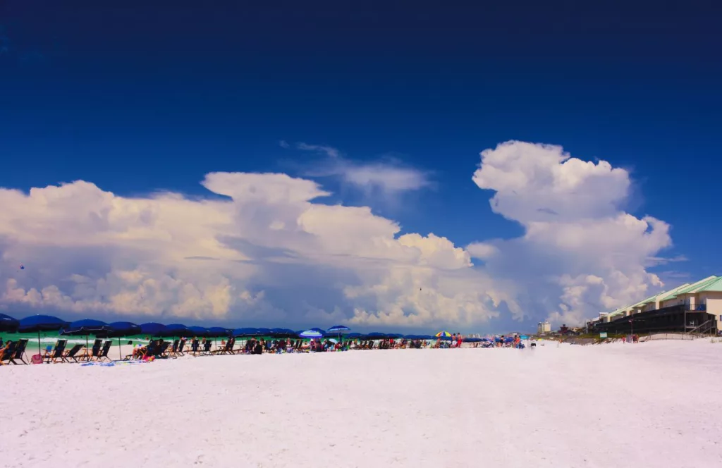 John Beasley Park with a bright blue sky and fluffy white clouds while people lay out on sun loungers with beach umbrellas. Keep reading to learn about the Best Public Beaches in Destin, Florida. 