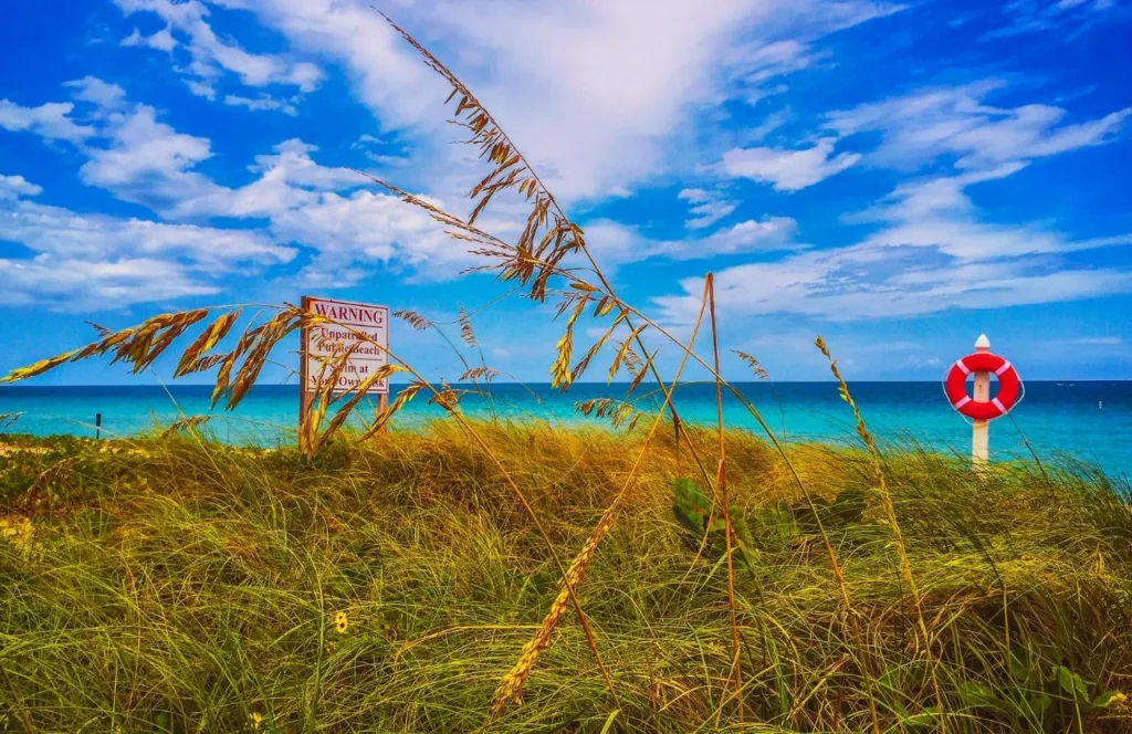 Sand dunes with a beach sign and bright blue waters at the beach in Florida. Keep reading to find what Florida beaches are the best to visit in September. 