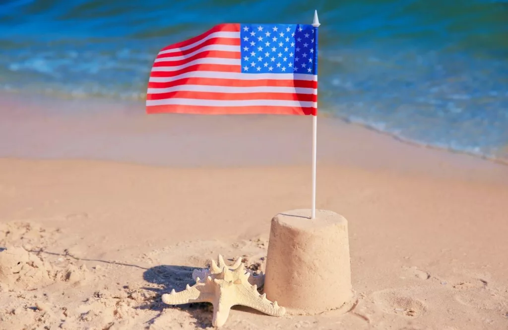 American flag in the top of a sand castle on the shore of the beach along side a big shell. Keep reading to learn more about Panama City, Florida snorkeling.